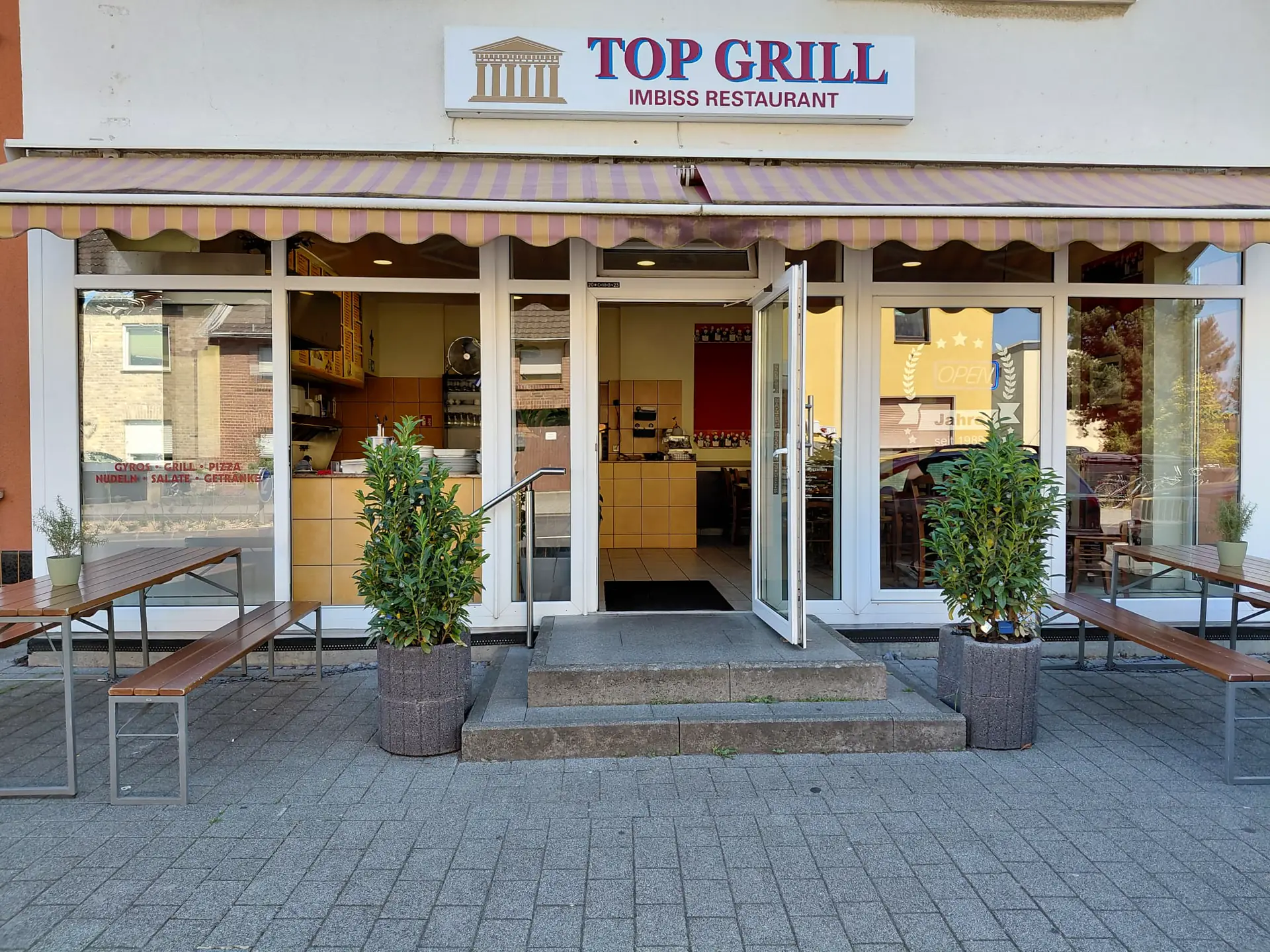 Ladenfront von Top Grill mit Schild, Markise, Bänken und offener Tür